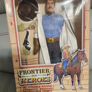 Puede incluir: Figura de acci&oacute;n de Jesse James de 30 cm de Frontier Heroes en caja. La figura viste camisa azul, vaqueros y abrigo blanco. Accesorios: sombrero, pistola y botas. La caja muestra un dibujo de un vaquero a caballo.