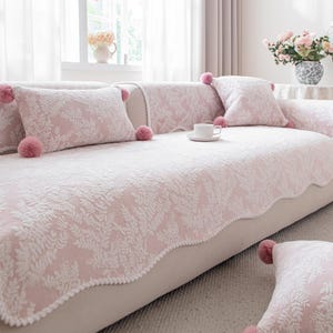 May include: A pale pink sofa cover and cushions with a white floral design and pink pom-poms. The sofa cover has a scalloped edge. A white teacup sits on the sofa. The room has a window with sheer curtains.