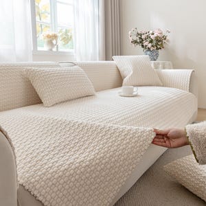 Może przedstawiać: Kremowa narzuta na sofę z teksturowanym, tkanym wzorem. Pokrycie jest narzucone na sofę, z pasującymi poduszkami. Ręka jest pokazana podczas regulacji pokrycia. Pokój ma jasną, przewiewną atmosferę z naturalnym światłem.