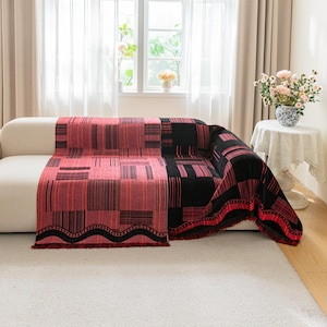 May include: A red and black patterned throw blanket draped over a white sofa. The blanket features a geometric design with rectangular and striped patterns. The fringe along the edge adds a decorative touch. The room has a window and a small table.