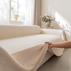 May include: Cream-colored sofa cover with a textured, speckled design. The cover is being lifted to reveal the sofa underneath. The room has a window with sheer curtains and a vase of flowers on a small table.