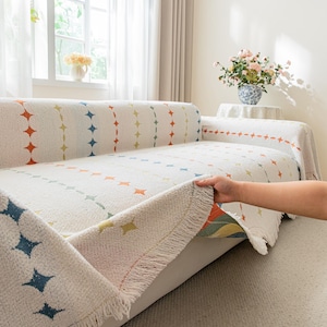 May include: Cream-colored sofa cover with a geometric pattern of colorful diamonds and lines. The cover has a fringed edge and is being held up to show the underside. The sofa is in front of a window with sheer curtains.