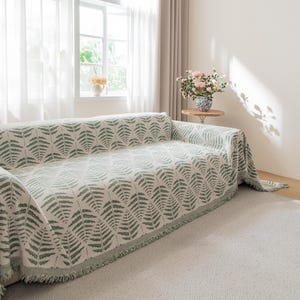 Può includere: Un divano color crema è coperto da una coperta con motivo a felci verdi. La coperta ha un bordo con frange e copre il divano. La stanza ha una finestra con tende trasparenti e un tavolino con fiori.
