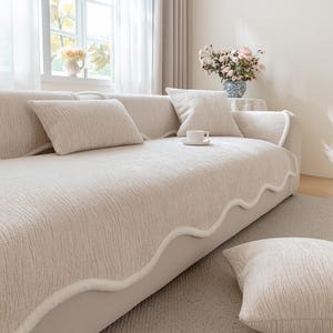 Könnte beinhalten: Beiges Sofa mit passenden Kissen. Das Sofa hat einen strukturierten Stoff und eine dekorative weiße Zierleiste an den Kanten. Eine weiße Tasse mit Untertasse steht auf dem Sofa. Der Raum hat ein Fenster mit transparenten Vorhängen.