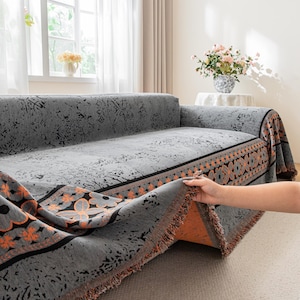 May include: Grey sofa cover with an orange and black floral border. The cover is draped over a grey sofa in a living room setting. The cover has a textured pattern and fringed edges.