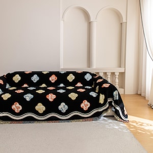 Könnte beinhalten: Eine schwarze Sofadecke mit einem sich wiederholenden Blumenmuster in Gelb, Orange und Blau. Die Decke hat einen weißen Wellenrand und Fransen am unteren Rand. Das Sofa steht auf einem weißen Teppich.