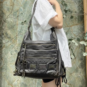 Può includere: Una borsa a tracolla in pelle nera con più scomparti con cerniera e borchie decorative in metallo. La borsa ha un aspetto invecchiato e una lunga tracolla regolabile. La borsa è indossata a spalla.