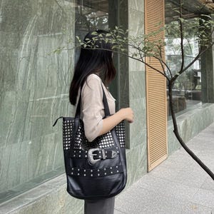 Puede incluir: Bolso tote negro con detalles de tachuelas plateadas y una hebilla grande. El bolso tiene forma rectangular y una correa larga para el hombro. El bolso lo lleva una persona.