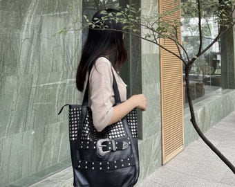 May include: Black tote bag with silver stud accents and a large buckle detail. The bag has a rectangular shape and a long shoulder strap. The bag is being carried by a person.