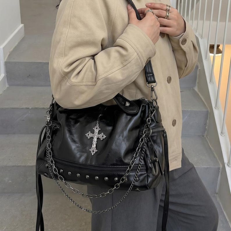 May include: Black faux leather shoulder bag with silver-tone cross pendant and chain accents. The bag features a top handle, a shoulder strap, and a zipper closure. Silver studs line the bottom of the bag.