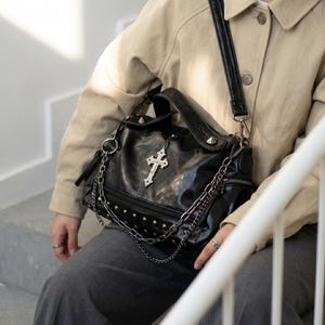 May include: Black leather handbag with silver-tone studs, chains, and a decorative cross pendant. The bag has a top handle and a shoulder strap. The bag is worn by a person wearing a tan jacket and gray pants.