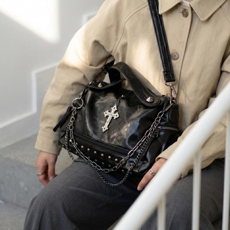 May include: Black leather handbag with silver-tone studs, chains, and a decorative cross pendant. The bag has a top handle and a shoulder strap. The bag is worn by a person wearing a tan jacket and gray pants.