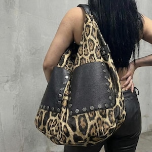 Puede incluir: Un bolso tote grande con estampado de leopardo y detalles en cuero negro. El bolso presenta bolsillos de cuero negro y está adornado con tachuelas plateadas. La correa también es de cuero negro.