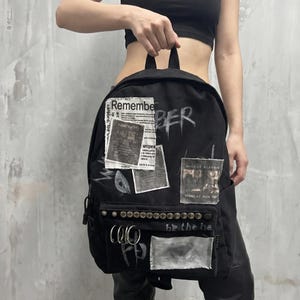 May include: Black backpack with multiple patches and silver studs. The backpack has the word "Remember" and "BER" painted on it. Patches include newspaper clippings and a small square with the text "MICHAEL ELAVIN".
