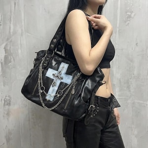 May include: Black leather handbag featuring a large cross design with a light blue background and silver chain accents. The bag has multiple compartments, zippers, and a shoulder strap. A stylish accessory.