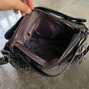 May include: Black shoulder bag with silver chain detailing and a cross charm. The bag has a top handle and a zippered main compartment. The interior is lined with a dark fabric. The bag is open, revealing the inside.