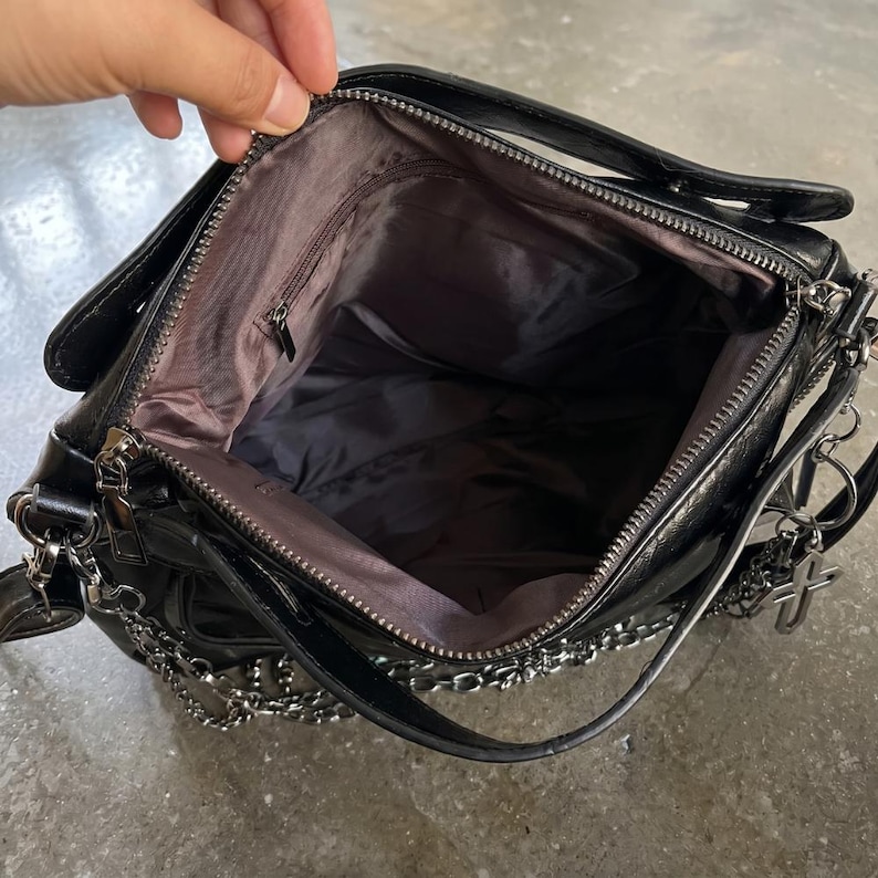 May include: Black shoulder bag with silver chain detailing and a cross charm. The bag has a top handle and a zippered main compartment. The interior is lined with a dark fabric. The bag is open, revealing the inside.