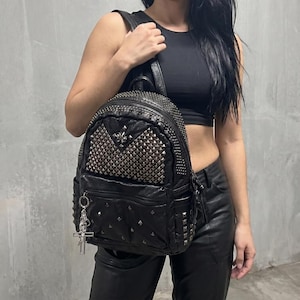 Könnte beinhalten: Schwarzer Lederrucksack mit silberfarbenen Nieten und einem Kreuzanhänger. Der Rucksack hat mehrere Taschen und ein Nietenmuster auf der Vorderseite. Er hat einen oberen Griff und verstellbare Schultergurte.