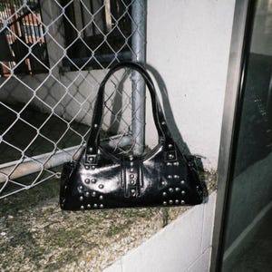 May include: A black handbag with silver-colored studs. The bag has two handles and a glossy finish. The bag is sitting on a ledge next to a chain-link fence.