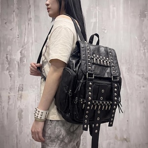 May include: A black backpack featuring a textured finish, adorned with silver-toned metal studs and rows of pointed spikes on the front flap and lower panel. The bag includes adjustable straps, side pockets, and a top handle for carrying.