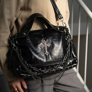 May include: Black handbag with a silver cross pendant and chain detailing. The bag features a textured finish, studded accents, and a shoulder strap. The design has a gothic or alternative aesthetic.