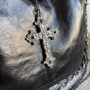 May include: Close-up of a black leather bag with a silver cross pendant. The cross is ornate with detailed filigree and hangs from a thin chain. The bag has a textured surface and a silver chain strap.
