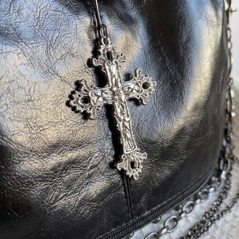 May include: Close-up of a black leather bag with a silver cross pendant. The cross is ornate with detailed filigree and hangs from a thin chain. The bag has a textured surface and a silver chain strap.