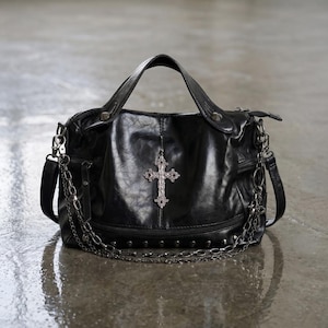 May include: Black handbag with a silver cross pendant. The bag has a top handle, a shoulder strap, and a chain detail. The bag is made of a textured material and has silver studs along the bottom.