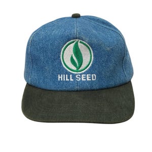 Pode incluir: Boné de beisebol azul denim com aba verde escuro. O boné apresenta um logotipo branco e verde com o texto "HILL SEED" bordado em branco. O boné tem um botão verde escuro na parte superior.
