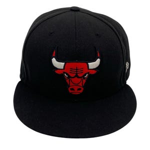May include: Black baseball cap featuring the Chicago Bulls logo. The logo is a red bull with white horns. The cap has a flat brim and a structured crown. A small logo is visible on the side.
