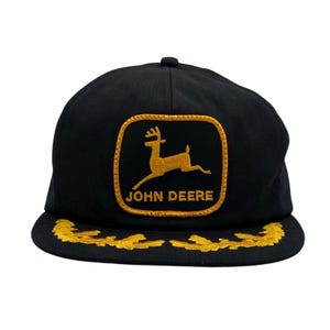 Pode incluir: Boné de beisebol preto com um patch bordado a ouro com o logotipo John Deere. O patch mostra um veado saltando. A aba tem detalhes bordados a ouro. O boné é um estilo clássico e ajustável.