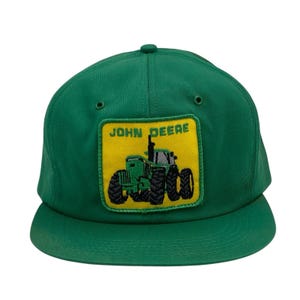 Op de afbeelding: Groene baseballcap met een John Deere tractor patch. De patch is een gele rechthoek met een groen tractorontwerp en de woorden "JOHN DEERE" in het zwart. De cap heeft een gebogen klep en een knoop aan de bovenkant.
