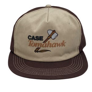 May include: A brown and tan baseball cap with the words "CASE Tomahawk" and a tomahawk graphic. The cap has a curved brim and a button on top. The stitching is white.