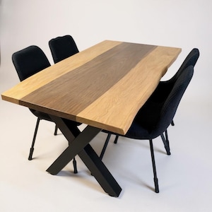Peut inclure: Une table à manger rectangulaire en bois avec un fini grain de bois naturel et une bande centrale marron foncé. La table a des pieds en métal noir et est entourée de quatre chaises en tissu noir.