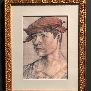 May include: A framed portrait of a person wearing a reddish-brown hat with a circular embellishment. The artwork is in a gold ornate frame with a white matting. The portrait is a pencil or charcoal drawing.