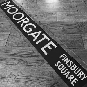 Puede incluir: Letrero rectangular negro con la palabra "MOORGATE" en letras mayúsculas blancas grandes. Debajo, el letrero dice "FINSBURY SQUARE" en letras mayúsculas blancas. El letrero está sobre un suelo de madera.