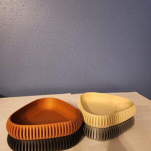 May include: Three triangular shaped dishes, one brown, one beige, and one gray, are stacked on a white surface. The dishes have a textured, ribbed design.