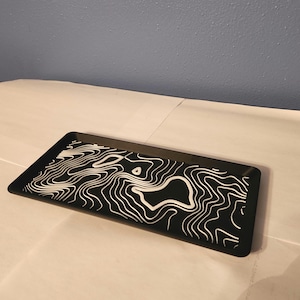 Peut inclure: Un plateau rectangulaire noir avec un motif de carte topographique blanc. Le plateau a un bord surélevé et une surface lisse. Le motif présente des lignes et des formes tourbillonnantes, créant un motif unique. Idéal pour servir ou exposer.