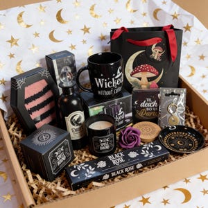 May include: A collection of witch-themed items in a gift box. Features a black mug with the text "Wicked without coffee", a black gift bag with a mushroom design, a coffin-shaped box, a candle, and black rose incense.