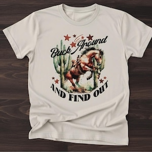 May include: Off-white t-shirt featuring a graphic of a bucking horse with a rider, cacti, and stars. The text "Buck Around and Find Out" is printed above and below the horse illustration.