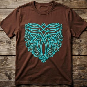 May include: A brown t-shirt featuring a teal, symmetrical design. The design is an intricate pattern of curved lines and floral elements, centered on the shirt's front. The shirt is displayed on a wooden surface.