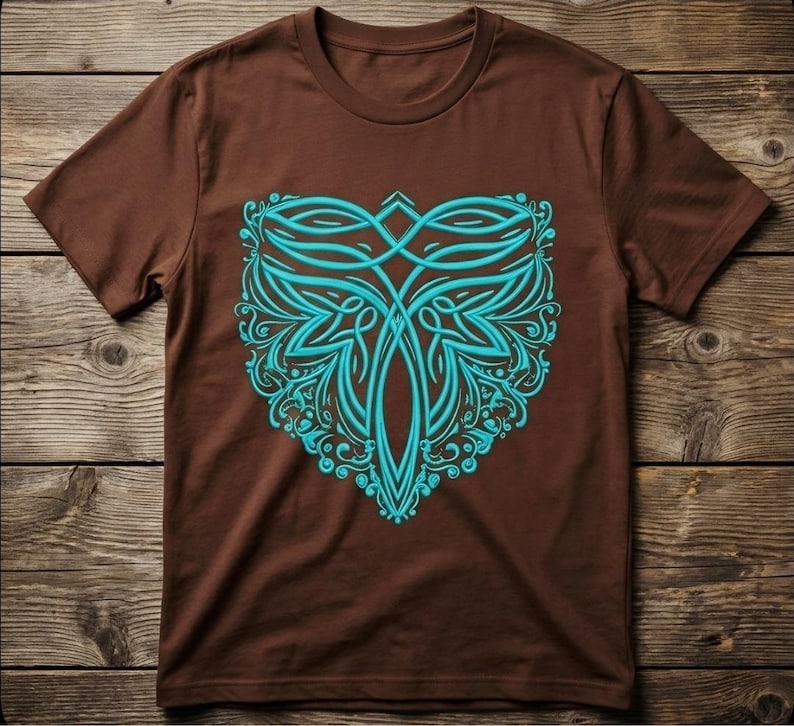 May include: A brown t-shirt featuring a teal, symmetrical design. The design is an intricate pattern of curved lines and floral elements, centered on the shirt's front. The shirt is displayed on a wooden surface.