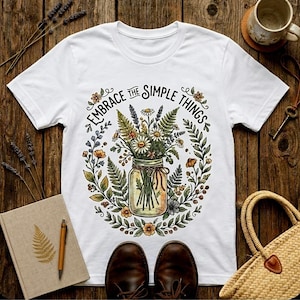 Op de afbeelding: Wit T-shirt met de tekst "Embrace the Simple Things" boven een bloemenillustratie van een pot gevuld met wilde bloemen. Het ontwerp bevat madeliefjes, varens en ander groen. Het shirt ligt op een houten oppervlak.
