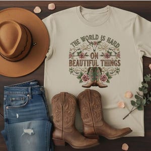May include: A tan t-shirt with the text "The World Is Hard On Beautiful Things" and a graphic of cowboy boots and roses. The image also includes a brown cowboy hat, blue jeans, and brown cowboy boots.