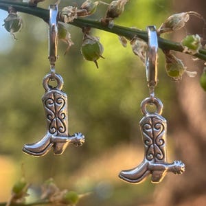 May include: A pair of silver-toned hoop earrings featuring dangling charms shaped like cowboy boots. Each boot charm displays intricate embossed scrollwork patterns and a small spur detail on the heel, suspended from a simple circular hoop closure.