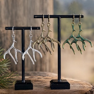 May include: Three pairs of dangling earrings shaped like deer antlers, displayed on two black T-shaped stands. One pair is polished silver, one is antique brass, and one is a weathered green patina finish, each featuring a secure lever-back closure.