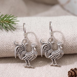 May include: A pair of silver-toned hoop earrings featuring dangling rooster charms. Each charm displays detailed engraved lines representing the feathers, comb, and tail of the bird. The earrings have a polished metallic finish and a hinged closure.
