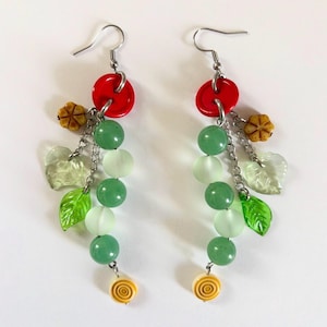 May include: A pair of dangling earrings with a nature-inspired design. Each earring features a red circular bead, green and white beads, clear leaf-shaped charms, and small yellow flower and spiral charms. The earrings are connected by silver-colored chains and hooks.
