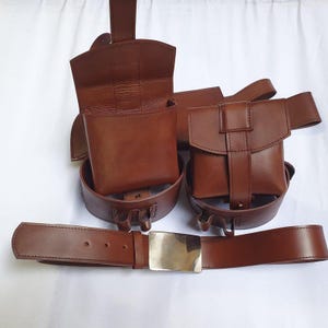 Puede incluir: Un cintur&oacute;n de cuero marr&oacute;n con hebilla plateada, acompa&ntilde;ado de dos bolsas a juego y una m&aacute;s grande, todas unidas al cintur&oacute;n. Las bolsas tienen forma rectangular con cierre de solapa. El cuero tiene una textura suave.