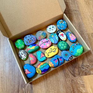 May include: A cardboard box filled with twenty smooth, hand-painted rocks featuring colorful designs including animals, insects, flowers, and inspirational text such as Cool to be Kind, dream, smile, and explore, all arranged in a collection.